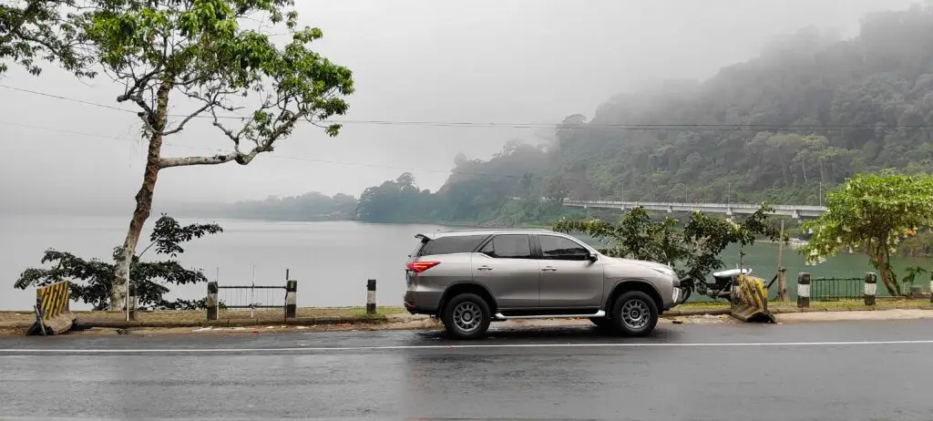 Rent Toyota Fortuner with Driveinbali.com – Driveinbali.com A gray Fortuner SUV is parked on a wet road next to a Bedugul Danau Beratan lake. Trees and foliage line the road, and a foggy forested hillside rises in the background, partially obscured by mist. Great place for the Toyota Fortuner!
